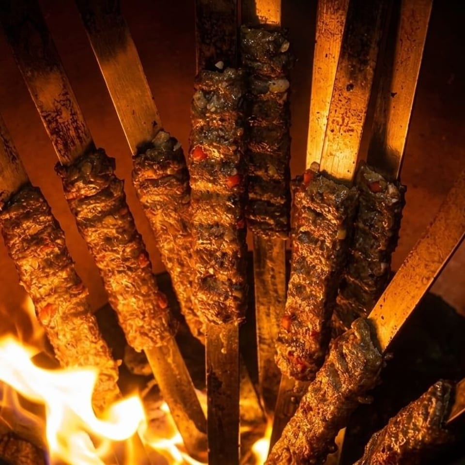 Skewered kebabs cooking over an open flame in a tandoor oven, glowing orange.