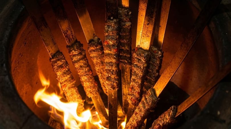 Skewered kebabs cooking over an open flame in a tandoor oven, glowing orange.