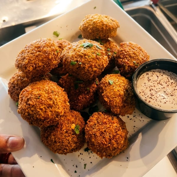 A hand holding a white plate piled high with golden-brown falafel balls and a side of white dipping sauce.