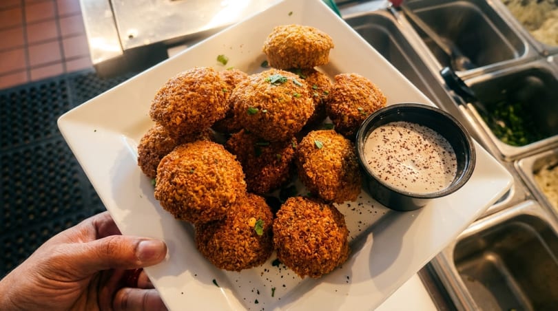 A hand holding a white plate piled high with golden-brown falafel balls and a side of white dipping sauce.